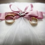 Close-up of elegant wedding rings on a silk pillow with a pink ribbon and a heart decoration, symbolizing love.